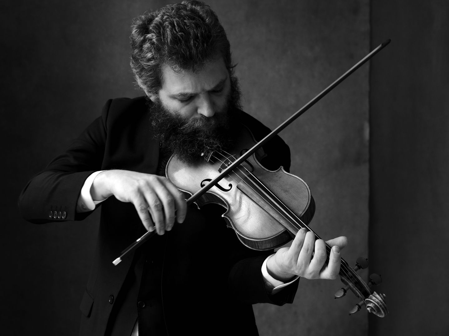 A man with a bushy beard, dressed in all black, plays a violin.